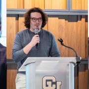 A three-panel collage of an event at the CU Denver Business School. The left panel shows a person viewing a research poster next to a large cassette tape graphic. The middle panel shows a young man speaking at a podium with the CU Denver Business School logo. The right panel shows two attendees networking with drinks and food in front of a research poster.