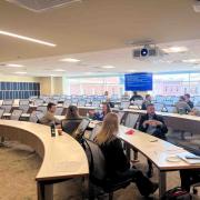 n interior view of a wide, sunlit classroom with tiered, curved wooden desks and grey ergonomic chairs. Participants are gathered in small clusters talking, while a blue presentation screen in the background displays discussion prompts titled "Layer 1: What Went Wrong, and When?" Large windows offer a view of downtown Denver buildings.