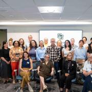 Group photo of the CU Denver Health Research Collaborative kickoff, with approximately two dozen participants gathered in a conference room, standing and seated in front of a screen displaying the CU Denver logo.