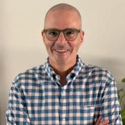 Hani Mansour smiles at camera. He is wearing a blue and white plaid button up shirt, and green rimmed glasses.