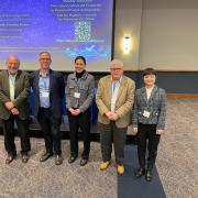 A group of five professionals stands in a conference room in front of a large screen displaying details for a "Generative AI Frontier" event in 2026. The group consists of three men and two women, dressed in business casual and professional attire, all wearing blue lanyards with name tags.  The man second from the left is David Hildebrand. He is wearing a navy blue blazer over a light blue button-down shirt and dark trousers, smiling at the camera.