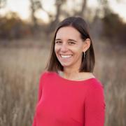 Liz D. standing outdoors in a field of tall grass, wearing a bright red three-quarter sleeve dress. She has straight, shoulder-length brown hair and is facing the camera, with trees softly blurred in the background and warm natural light.