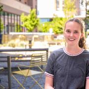 Martha smiles at the camera, wearing a grey tshirt. The background is a couryard with blooming green trees and city building.
