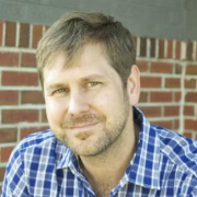 Wayne Miller, with short light-brown hair and a trimmed beard, is sitting in front of a brick wall. He is wearing a blue plaid shirt and looking at the camera with a slight smile