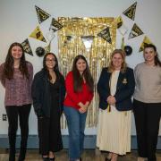 Five CLAS Outstanding Graduate Award recipients stand together in front of a gold graduation-themed backdrop, smiling during the Fall 2025 recognition event.