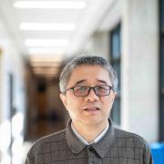  Dr. Haobin Wang, Professor of Chemistry at the University of Colorado Denver, poses in a bright university corridor. He is wearing glasses and a gray plaid polo shirt, with a softly blurred hallway visible behind him