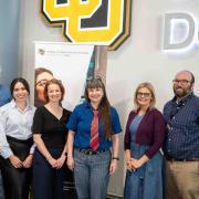 From left to right Jim Grigsby, Vanessa Navarro-Rodríguez, Anna Warrener, Nicky Beer, Lisa Keranen, and Michael Moore. They stand in front of a University of Colorado Denver sign and a banner for the College of Liberal Arts and Sciences 