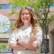 Kayley stands smiling with arms crossed in front of clock