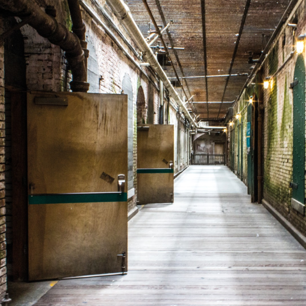 Brick hallway with pipes running along the ceiling. Wooden doors are open on the left and green metal doors are closed on the right. Sign on the right reads "Alcatraz". 