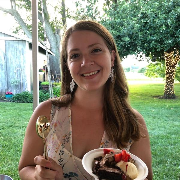 Portrait of Angie Schmitt. Outdoors and smiling, she holds a gold spoon in her left hand and a bowl full of ice cream, brownies, and cherries. 