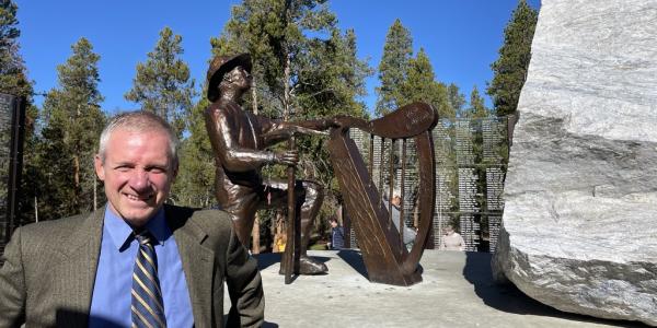 Jim Walsh smiling in front of memorial statue of a miner kneeling in front of a Celtic harp