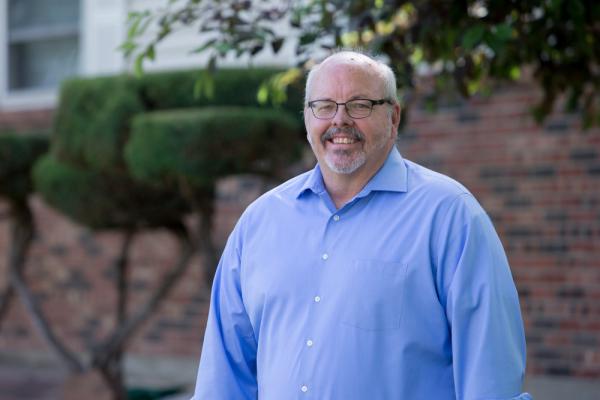 Photo of Tom Sullivan in blue shirt looking directly into the camera and smiling wearing glasses