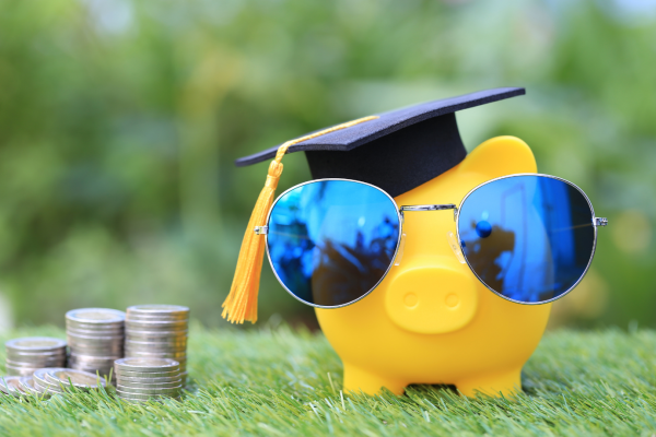 A piggy bank wearing sunglasses and a graduation cap with a pile of quarters next to it