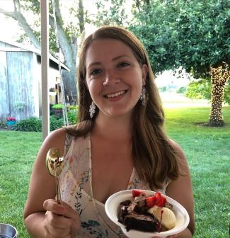Portrait of Angie Schmitt. Outdoors and smiling, she holds a gold spoon in her left hand and a bowl full of ice cream, brownies, and cherries.