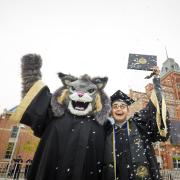 Picture of Milo the Lynx mascot in a graduation gown with a student in a graduation cap and gown, in front of the red Tivoli building.