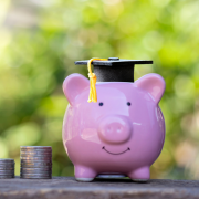 Photo of 3 stacks of coins sitting next to a piggy bank with a graduation cap on