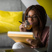 A woman sits in front of a grey seat sipping tea while contemplating her writing on the notebook in front of her.