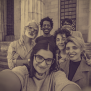 A group of students and faculty member take a smiling selfie in front of a building with two columns.
