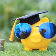 A piggy bank wearing sunglasses and a graduation cap with a pile of quarters next to it
