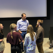 Phil Weiser and various students speaking in a dark room with a white screen in the background