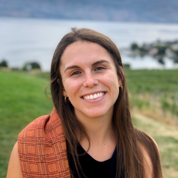 Headshot of Emilia with brown hair, an orange sweater over a black shirt, with grass and water in the background