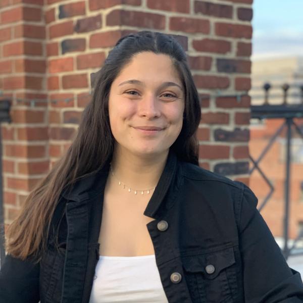 Fiona with brown hair, black denim jacket over white shirt against brick wall