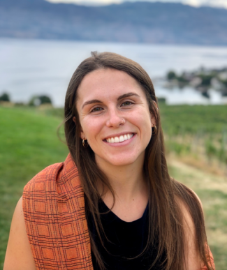 Headshot of Emilia with brown hair, an orange sweater over a black shirt, with grass and water in the background