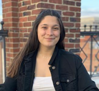 Fiona with brown hair, black denim jacket over white shirt against brick wall